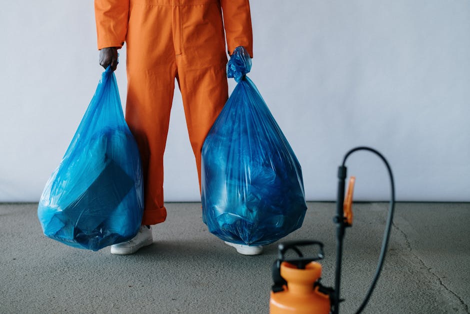 A person wearing orange overalls holding two large blue garbage bags filled with waste, standing on a grey carpeted floor in a room with plain white walls. The individual's footwear is visible, with w