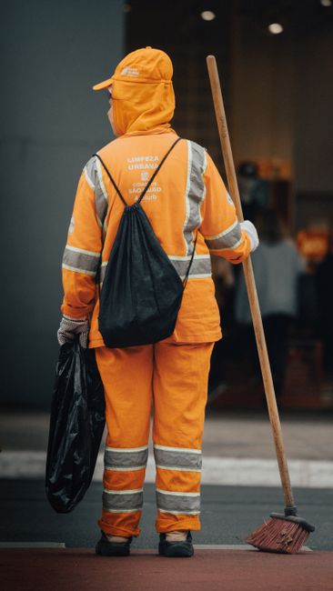 A person wearing orange overalls holding two large blue garbage bags filled with waste, standing on a grey carpeted floor in a room with plain white walls. The individual's footwear is visible, with w